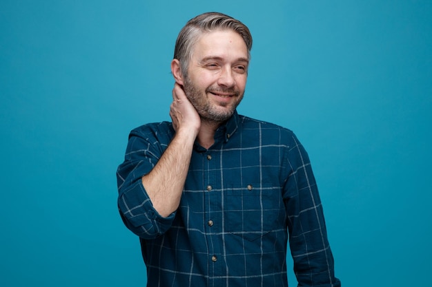 Hombre de mediana edad con cabello gris en camisa de color oscuro mirando a un lado sonriendo con cara feliz tocando su cuello de pie sobre fondo azul