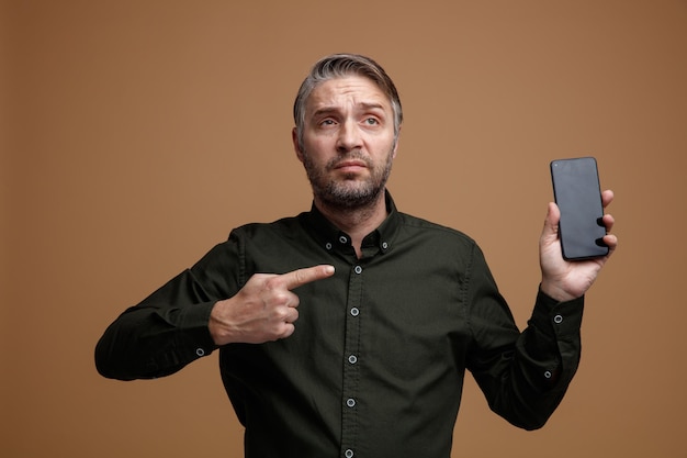Hombre de mediana edad con cabello gris en camisa de color oscuro que muestra el teléfono inteligente apuntando con el dedo índice decepcionado y triste de pie sobre fondo marrón