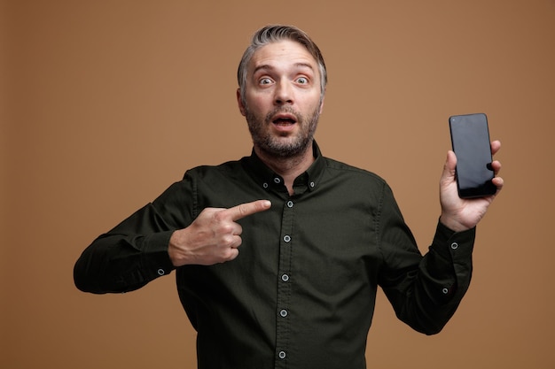 Hombre de mediana edad con cabello gris en camisa de color oscuro que muestra el teléfono inteligente apuntando con el dedo índice mirando a la cámara asombrado y sorprendido de pie sobre fondo marrón