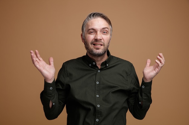 Hombre de mediana edad con cabello gris en camisa de color oscuro que parece confundido levantando los brazos sin respuesta de pie sobre fondo marrón