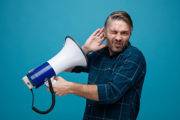 Foto hombre de mediana edad con cabello gris en camisa de color oscuro sosteniendo un megáfono cerca de su oído luciendo confundido y disgustado de pie sobre fondo azul