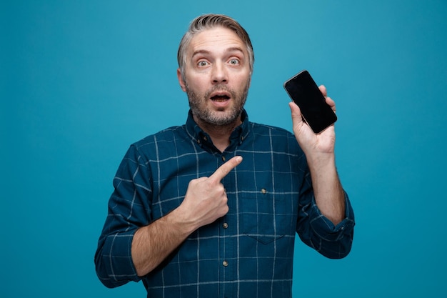 Hombre de mediana edad con cabello gris en camisa de color oscuro sosteniendo un teléfono inteligente apuntando con el dedo índice mirando sorprendido y asombrado de pie sobre fondo azul