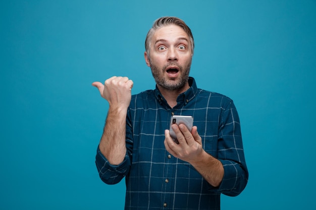 Hombre de mediana edad con cabello gris en camisa de color oscuro sosteniendo un teléfono inteligente apuntando con el pulgar hacia un lado mirando a la cámara asombrado y sorprendido de pie sobre fondo azul