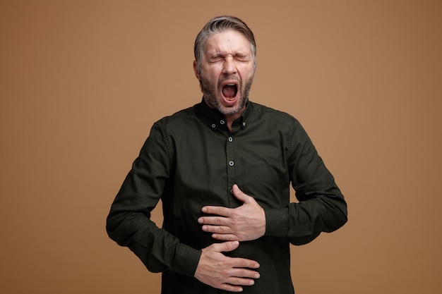 Hombre de mediana edad con cabello gris en camisa verde oscuro que parece cansado bostezando quiere dormir tomados de la mano en su vientre de pie sobre fondo marrón