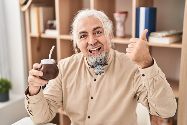 Hombre de mediana edad con canas bebiendo infusión de mate apuntando con el pulgar hacia el lado sonriendo feliz con la boca abierta