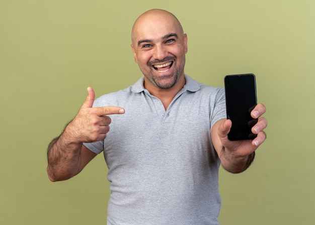 Hombre de mediana edad casual alegre que muestra el teléfono móvil apuntando a él aislado en la pared verde oliva