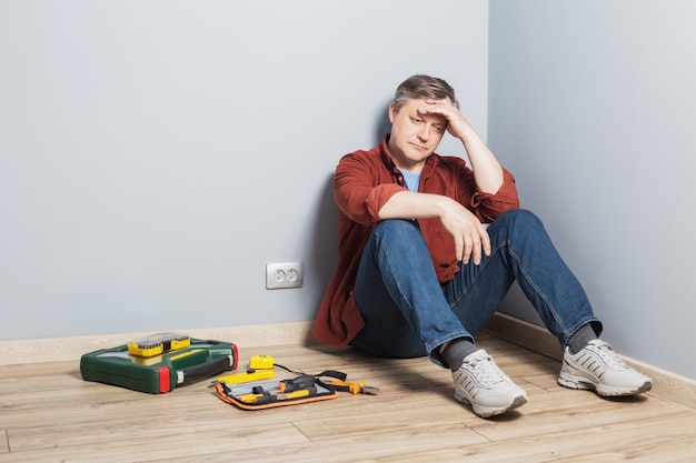 Hombre de mediana edad descansando sentados en el suelo junto a una caja de herramientas de reparación