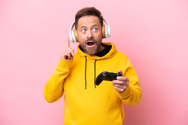 Hombre de mediana edad jugando con un controlador de videojuegos aislado en un fondo rosa pensando en una idea apuntando con el dedo hacia arriba