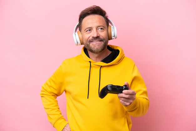 Hombre de mediana edad jugando con un controlador de videojuegos aislado en un fondo rosa posando con los brazos en la cadera y sonriendo