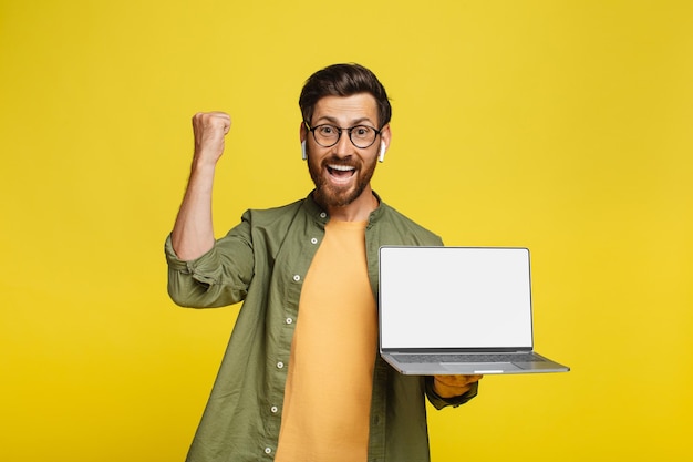 Foto hombre de mediana edad lleno de alegría sosteniendo una computadora portátil con pantalla en blanco y agitando los puños apretados maqueta de fondo amarillo