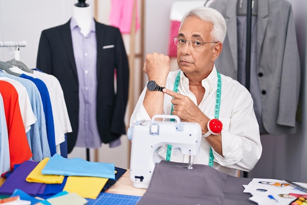 Hombre de mediana edad con una modista de pelo gris que usa una máquina de coser con prisa apuntando a ver la impaciencia del tiempo mirando a la cámara con una expresión relajada