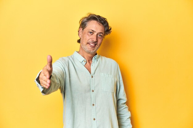 Hombre de mediana edad posando sobre un fondo amarillo estirando la mano a la cámara en gesto de saludo