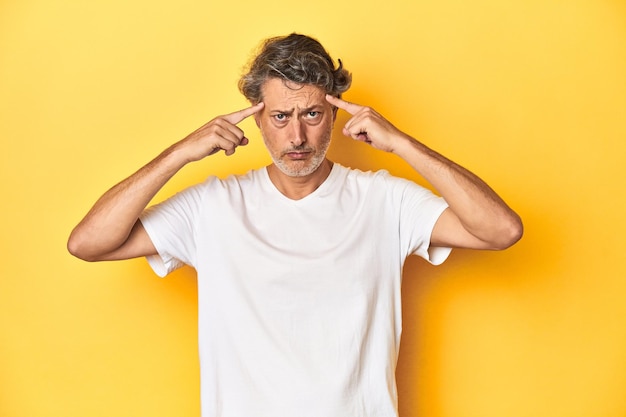 Hombre de mediana edad posando sobre un telón de fondo amarillo centrado en una tarea manteniendo los dedos índice apuntando a la cabeza