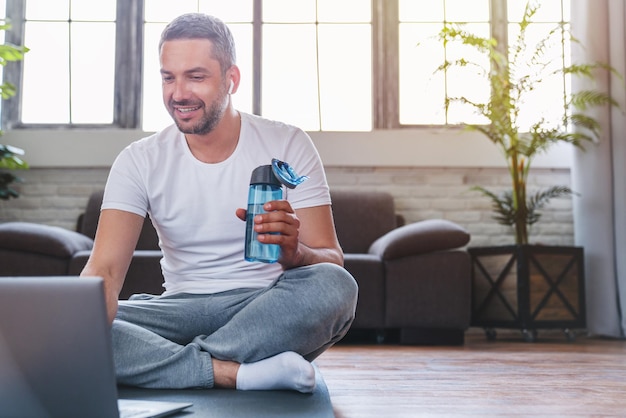 Hombre de mediana edad que usa una computadora portátil y bebe agua mientras toma un descanso durante el entrenamiento en casa