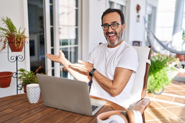 Hombre de mediana edad que usa computadora portátil en casa señalando a un lado con las manos abiertas mostrando espacio de copia, presentando un anuncio sonriendo emocionado feliz