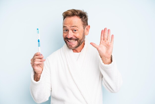 Hombre de mediana edad sonriendo felizmente, saludando con la mano, dándote la bienvenida y saludándote. concepto de cepillo de dientes