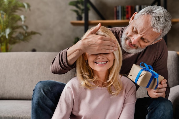 Hombre de mediana edad sorprendiendo con caja de regalo a su esposa cerrando los ojos