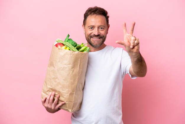 Hombre de mediana edad sosteniendo una bolsa de compras aislada en un fondo rosa feliz y contando tres con los dedos