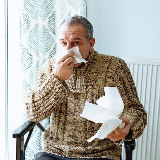 Hombre de mediana edad con suéter abrigado, se suena la nariz con una servilleta de papel desechable, estornuda y tose