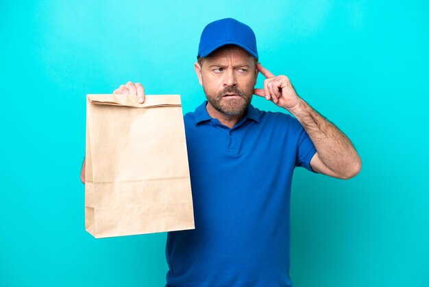 Hombre de mediana edad tomando una bolsa de comida para llevar aislada de fondo azul con dudas y pensando