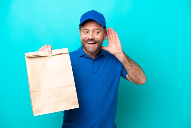 Hombre de mediana edad tomando una bolsa de comida para llevar aislada de fondo azul escuchando algo poniendo la mano en la oreja