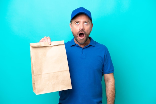 Hombre de mediana edad tomando una bolsa de comida para llevar aislada de fondo azul mirando hacia arriba y con expresión sorprendida