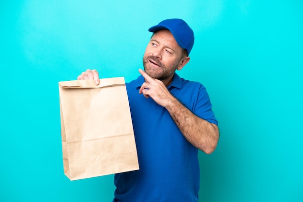 Hombre de mediana edad tomando una bolsa de comida para llevar aislada de fondo azul mirando hacia arriba mientras sonríe