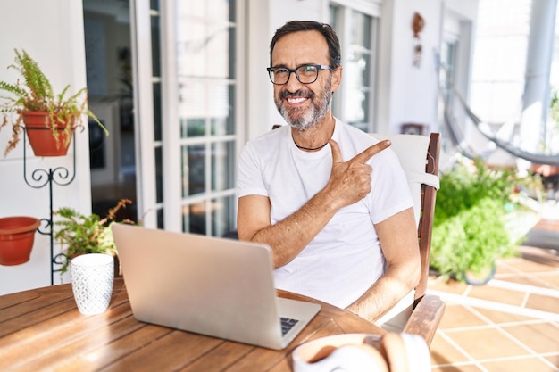 Hombre de mediana edad usando computadora portátil en casa alegre con una sonrisa en la cara apuntando con la mano y el dedo hacia un lado con expresión feliz y natural