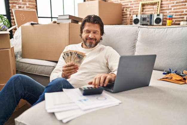 Hombre de mediana edad usando una computadora portátil contando billetes de coronas dinamarcas en su nuevo hogar