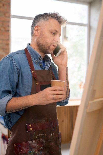 Hombre de mediana edad con un vaso de café de pie delante del caballete en el estudio mientras habla con alguien por teléfono móvil