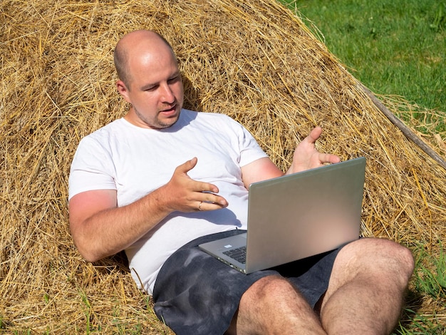 Un hombre de mediana edad vestido con una camiseta y pantalones cortos está acostado en un pajar amarillo con una computadora portátil gris en sus manos Entrenamiento de trabajo remoto Área rural Indignación en la emoción de la cara