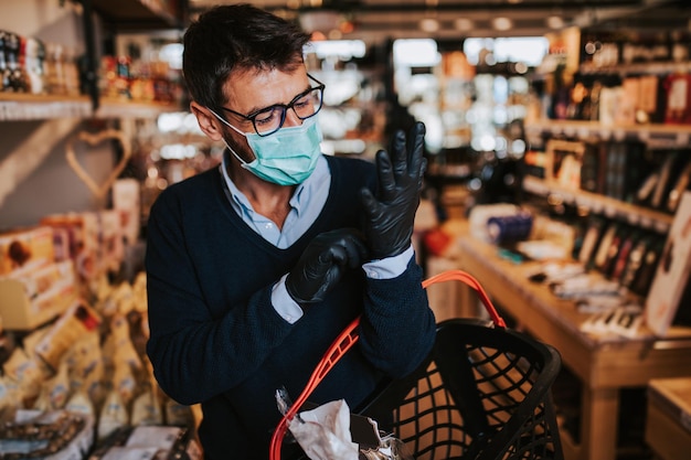 Hombre de mediana edad vestido informalmente con máscara protectora y guantes comprando alimentos y bebidas saludables en un moderno supermercado o tienda de abarrotes. Estilo de vida pandémico o epidémico.