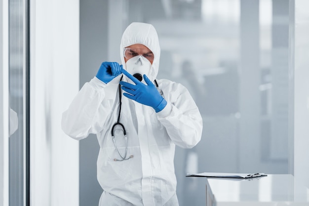 Hombre médico científico en bata de laboratorio, gafas defensivas y máscara quitándose guantes azules