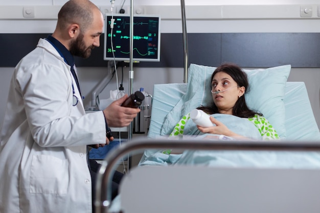 Hombre médico especialista explicando el tratamiento de las pastillas contra el dolor durante la cita clínica en la sala del hospital. Mujer enferma con tubo de oxígeno nasal sentada en la cama discutiendo medicamentos vitamínicos