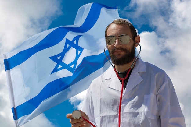 Hombre médico judío americano encantador barbudo en Yarmulke blanco (sombrero, Kippah, sombrero judío) con gafas de sol, abrigo y estetoscopio en el fondo de la bandera de Israel. Doble exposición de hombre guapo ruso