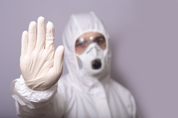 Hombre, médico en traje de protección, con máscara, gafas y guantes contra la infección, covid 19, durante una pandemia, se detiene con la mano, se detiene, se queda en casa.