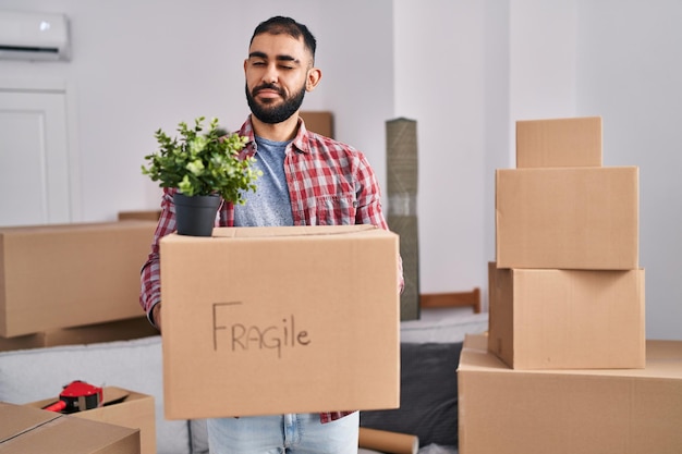 Hombre de Medio Oriente con barba mudándose a un nuevo hogar sosteniendo una caja de cartón guiñando un ojo mirando a la cámara con expresión sexy, cara alegre y feliz.