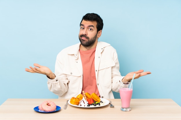 Hombre en una mesa desayunando gofres y un batido con dudas mientras levanta las manos