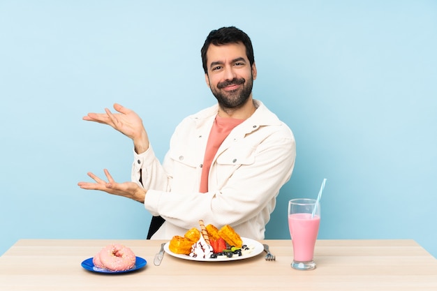 Hombre en una mesa desayunando gofres y un batido extendiendo las manos hacia un lado para invitar a venir