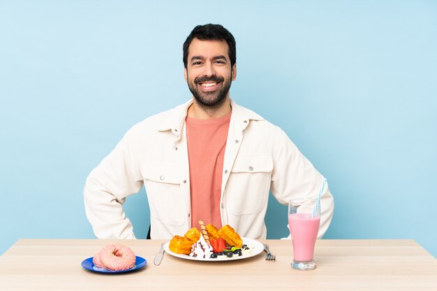 Hombre en una mesa desayunando gofres y un batido posando con los brazos en la cadera y sonriendo