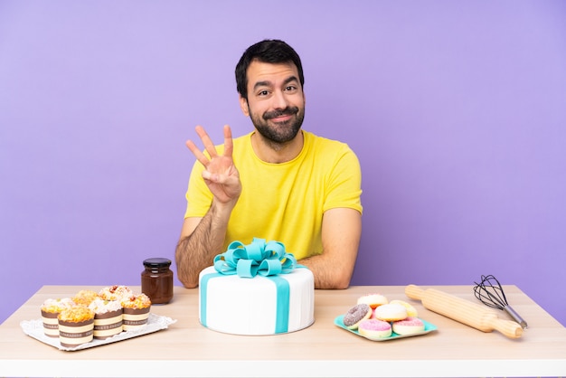 Hombre en una mesa con un gran pastel feliz y contando tres con los dedos