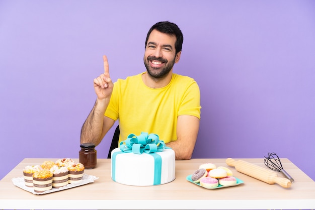 Hombre en una mesa con un gran pastel mostrando y levantando un dedo en señal de lo mejor