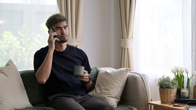 Hombre milenario casual hablando por teléfono móvil y bebiendo café en la sala de estar