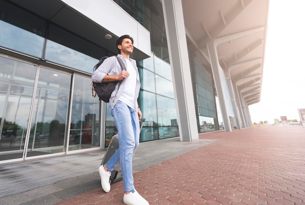 Hombre milenario feliz caminando con equipaje fuera del edificio del aeropuerto