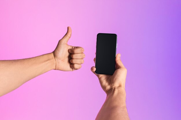 Hombre milenario irreconocible mostrando teléfono inteligente con pantalla vacía gesticulando con el pulgar hacia arriba en luz de neón