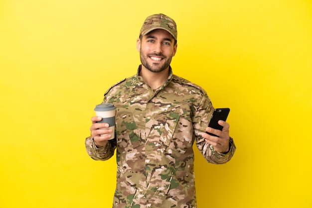 Hombre militar aislado sobre fondo amarillo con café para llevar y un móvil