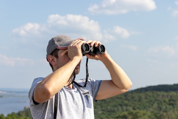 Un hombre mira a través de binoculares contra una vista panorámica de un ancho río en una colina en un día soleado