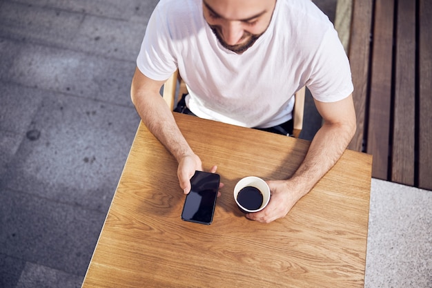 Hombre mirando al teléfono inteligente y sosteniendo café vista exterior desde arriba