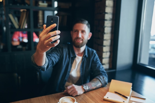 Hombre mirando a la cámara mientras hace una selfie. Hombre joven barbudo, vestido con una camisa de vaqueros, sentado en una mesa en un café y usando un teléfono inteligente