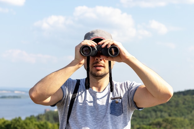 Un hombre mirando a la cámara a través de binoculares contra una vista panorámica de un ancho río en una colina en un día soleado